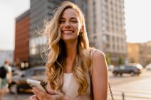 Urbana, nasmejana, raspoložena devojka šeta kroz grad sa telefonom u rukama