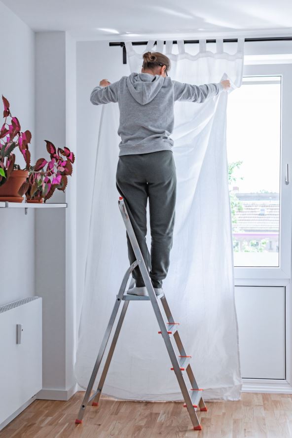 stockphotowomanstandsonastepladdernearthewindowandhangswhitecurtainsonthecurtainrod1499794904.jpg