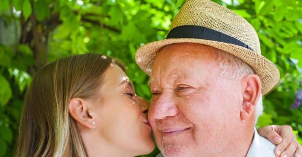 Stariji čovek sa šeširom nasmešen dok ga u obraz ljubi devojka