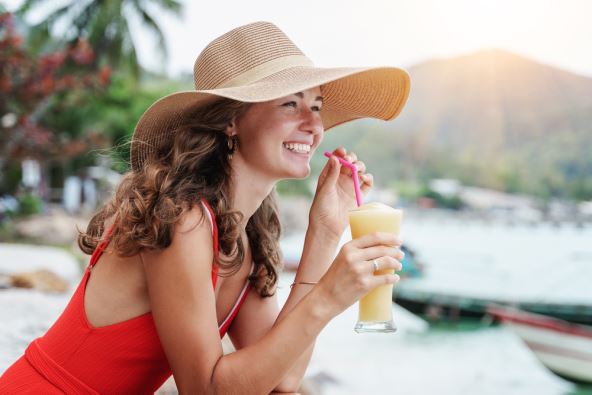 Žena u crvenom kupaćem kostimu i sa šeširom na plaži pije koktel
